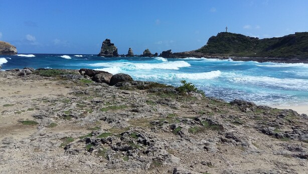 A rocky beach with waves crashing onto the shore, a cross on a hill, and a clear blue sky. - GÎTE LES PIMENTIERS DE PAPAYE (Saint Claude)
