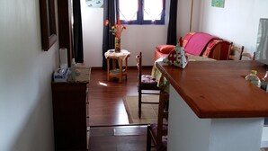 A cozy living area with wooden floors, a dining table, and a sofa. There is a window with curtains, a lamp, and a painting on the wall. - GÎTE LES PIMENTIERS DE PAPAYE (Saint Claude)