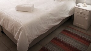 A neatly made bed with white linens, a red and gray striped rug, a white nightstand with a lamp, and a painting of a bridge on the wall.
