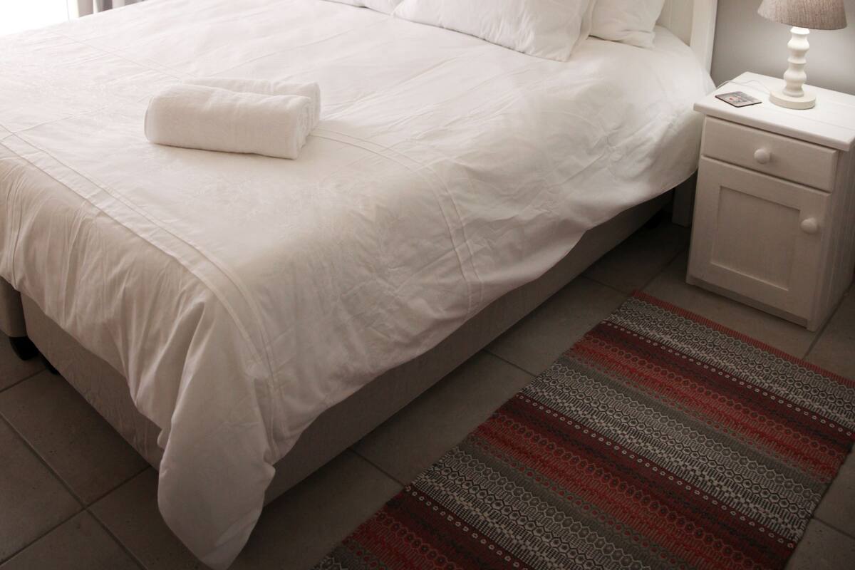 Una cama bien hecha con sábanas blancas, una alfombra a rayas grises y rojas, una mesita de noche blanca con una lámpara, y un cuadro de un puente en la pared.