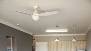 A modern living room with a ceiling fan, a kitchen area with wooden cabinets, and a dining area with white bar stools.