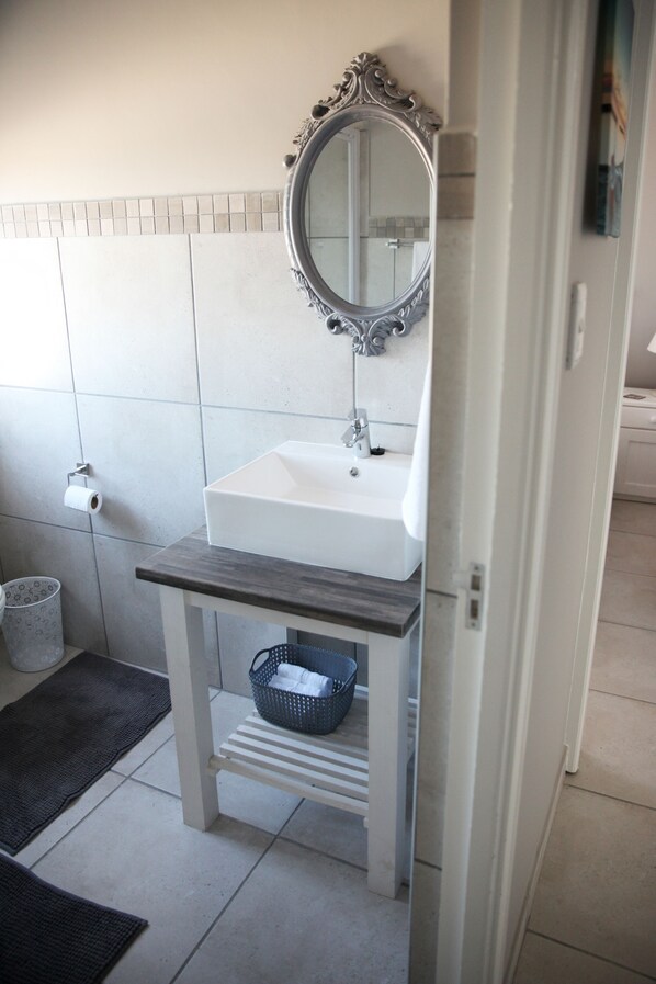 Un baño con un lavamanos blanco, un espejo con un marco decorativo, un perchero y un cubo de basura.
