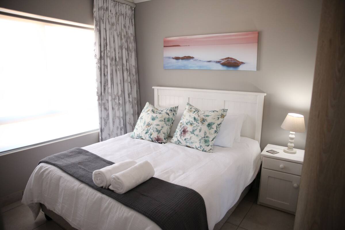 Una cama bien hecha con almohadas estampadas, un cabecero blanco y una mesita de noche con una lámpara.