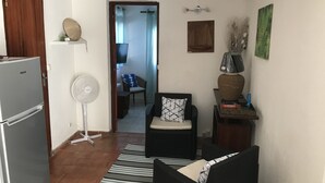 Een ruimtelijk ingerichte leefruimte met een koelkast, een ventilator, een bank, een stoel en een kleine tafel met decoratieve objecten.