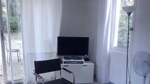 Una habitación pequeña y luminosa con una mesa blanca, una silla de oficina negra, un televisor de pantalla plana y un ventilador de pie.