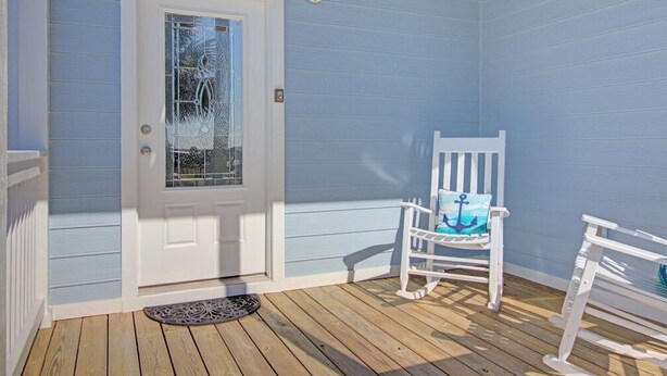 Eine Veranda mit einer weißen Tür, einem Schaukelstuhl mit blauer Sitzkissen und einer kleinen weißen Bank.