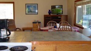 A kitchen with a stove, a coffee maker, a dining table with chairs, a television on a stand, and a window with blinds.