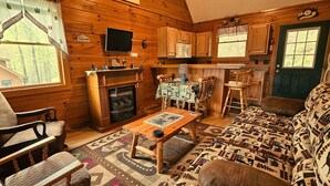 A cozy cabin interior with a fireplace, wooden furniture, a dining area, and a kitchen with wooden cabinets and a window. - Deer Country Vacation Cabins #4 - Long Range View, Dogs Friendly (Waynesville)