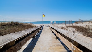 Ein hölzerner Steg führt zu einem Sandstrand, in der Ferne weht eine Flagge.
