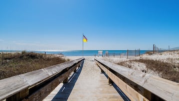 En træbro fører hen til en sandstrand, hvor et flag vinker i det fjerne.