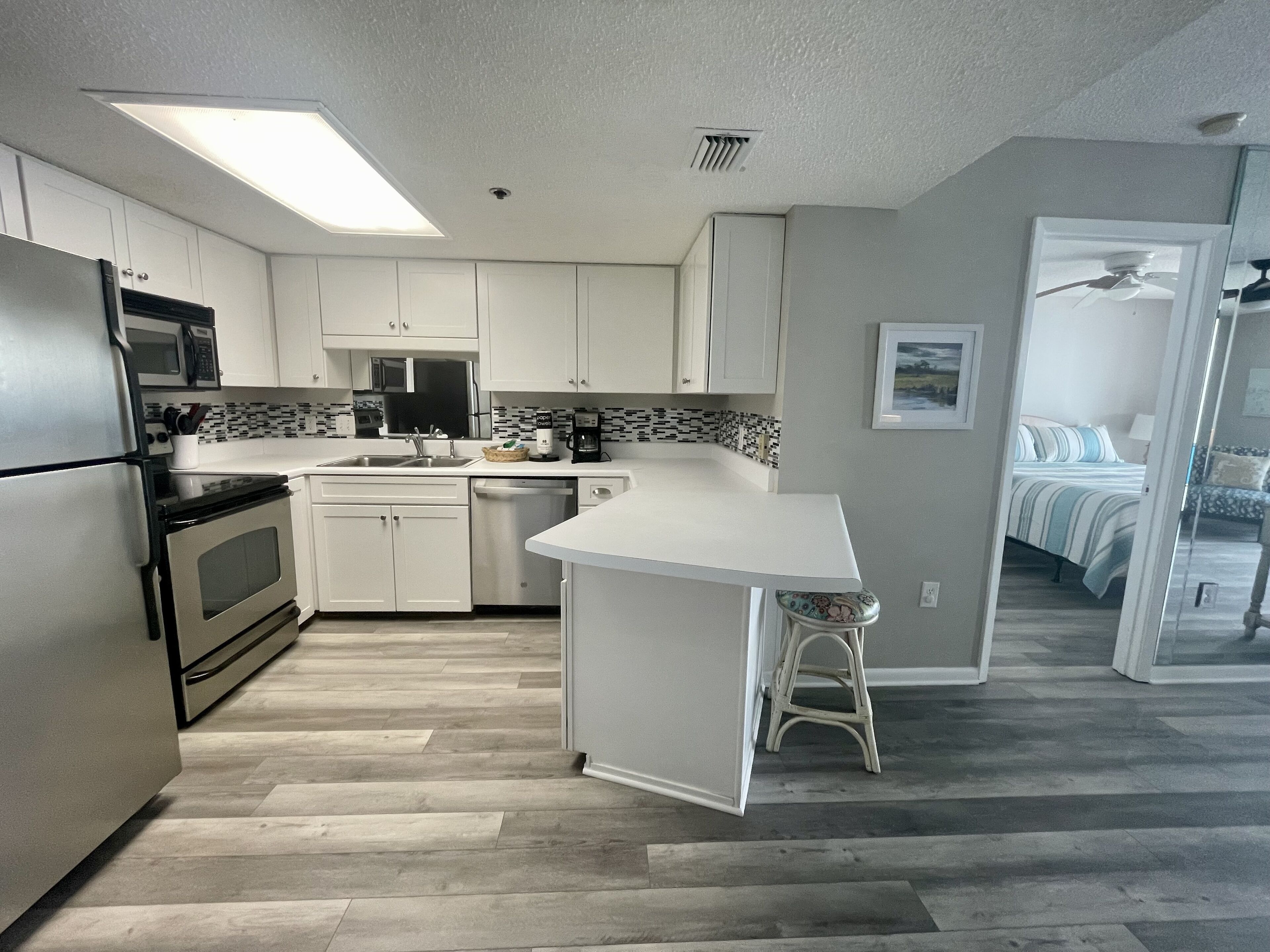 A modern kitchen with white cabinets, a stainless steel refrigerator, a microwave, and a coffee maker. There is a small island with a stool, and a view into a bedroom with a bed and a chair.