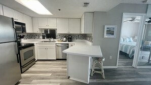 Eine moderne Küche mit weißen Schränken, einem Edelstahlkühlschrank, einer Mikrowelle und einer Kaffeemaschine. Eine kleine Kücheninsel mit einem Hocker, und ein Blick in ein Schlafzimmer mit Bett und Sessel.