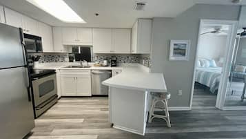 A modern kitchen with white cabinets, a stainless steel refrigerator, a microwave, and a coffee maker. There is a small island with a stool, and a view into a bedroom with a bed and a chair.