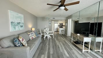 A modern living room with a grey sofa, white chairs, a wooden ceiling fan, and a kitchen area with a refrigerator and microwave.