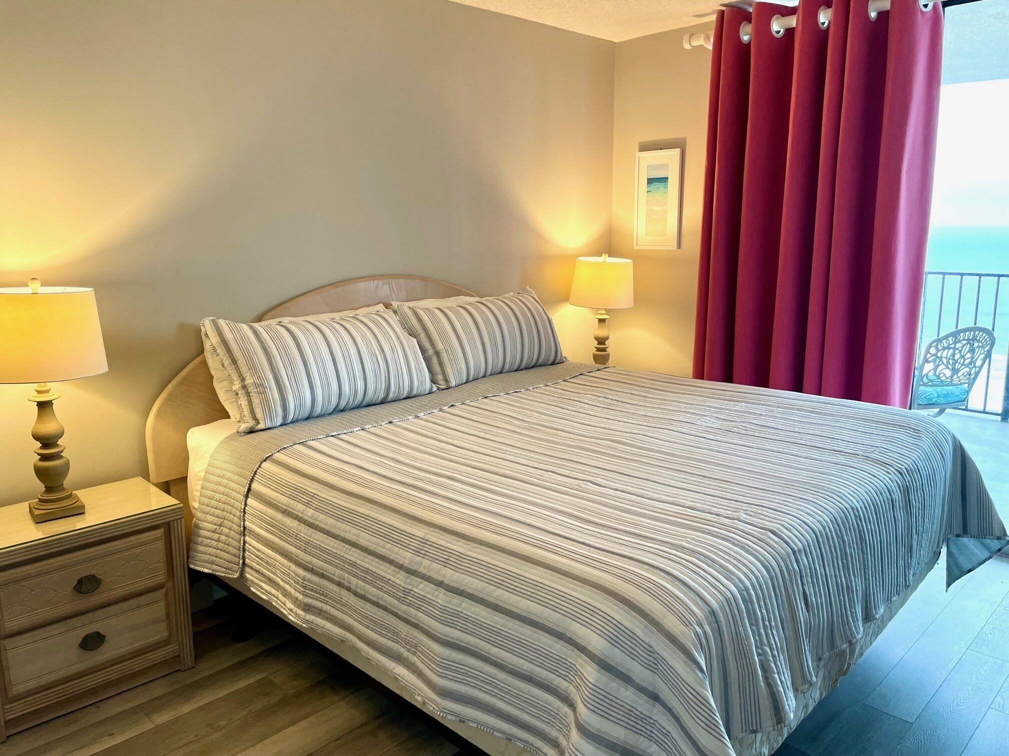 Cama bem arrumada com roupa de cama listrada, duas luminárias de cabeceira, uma mesa de cabeceira de madeira e vista para o mar por cortinas vermelhas.