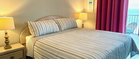 Cama bem arrumada com roupa de cama listrada, duas luminárias de cabeceira, uma mesa de cabeceira de madeira e vista para o mar por cortinas vermelhas.