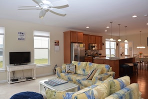 A living room with a sofa, a coffee table, and a TV on a stand. There is a ceiling fan, a kitchen area with cabinets and appliances, and a dining area with chairs and a table. - Beautiful Rehoboth Beach Townhouse w/ Pool Sleeps 8 (Rehoboth Beach)