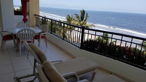 Un balcon avec vue sur l’océan, des chaises en plastique blanches, un parasol rouge et une table blanche.