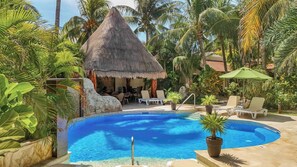 Una piscina rodeada de palmeras, una cabaña con techo de paja y sillas de descanso.