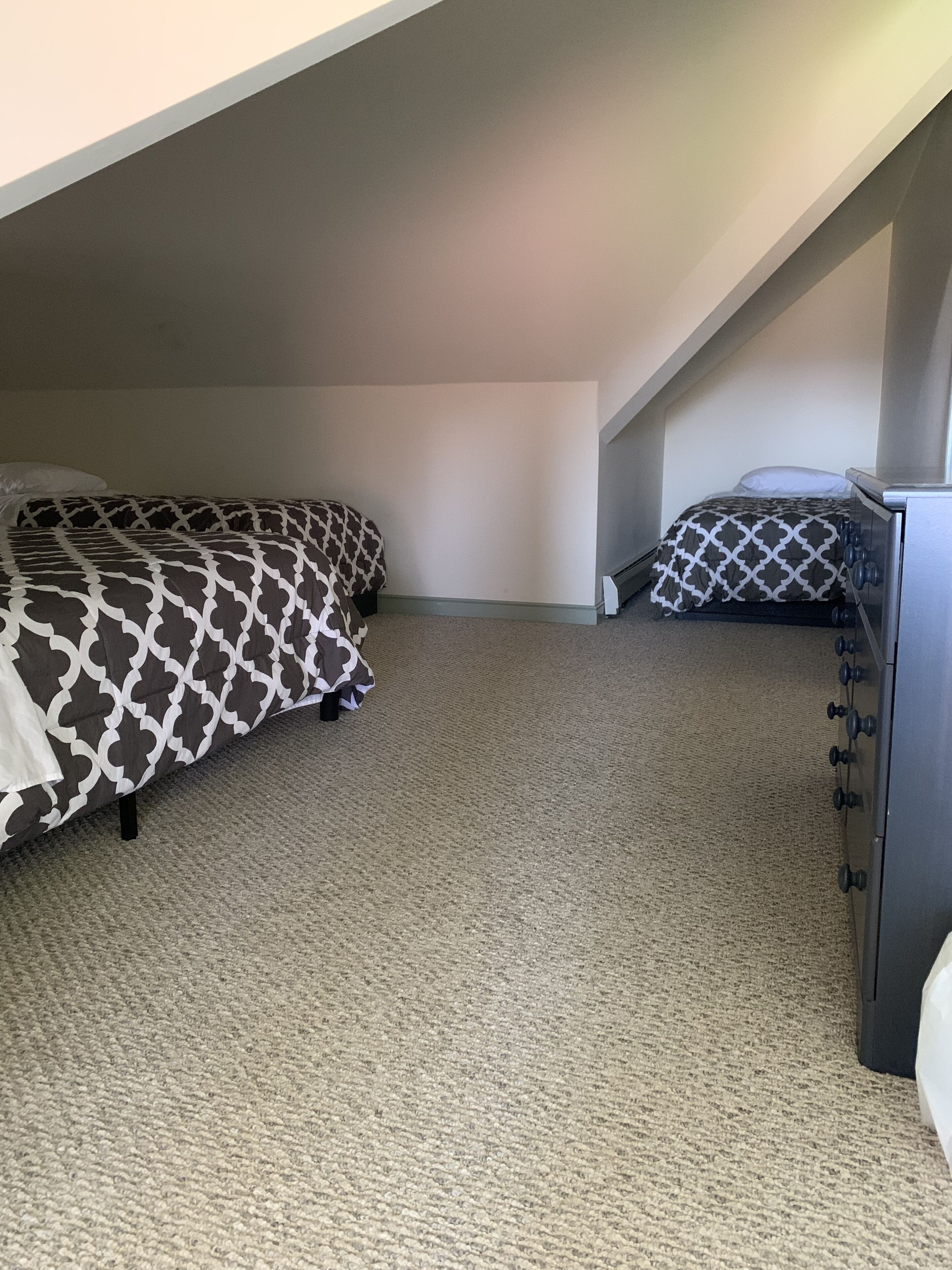 A small, dimly lit room with two beds, a dresser, and a sloped ceiling.