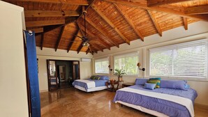 A spacious room with a high wooden ceiling, two beds with blue and white bedding, a small table with a plant, and a sliding glass door leading to another area. - Beautiful Tropical Island Villa for big families and groups (Frederiksted)