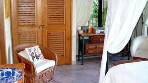 Una habitación con puerta de madera, una silla de mimbre con un cojín colorido, un aparador de madera y una cama con dosel blanco.