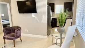 A modern living room with a flat-screen TV mounted on the wall, a glass dining table with chairs, a purple armchair, and a potted plant. - WEEKLY or MONTH TO MONTH LUXURY VACATION RENTAL - AP1  (Pompano Beach)