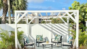 A covered outdoor seating area with chairs and a table, surrounded by plants and a white fence. - WEEKLY or MONTH TO MONTH LUXURY VACATION RENTAL - AP1  (Pompano Beach)