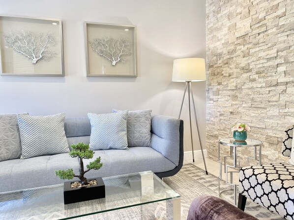 A modern living room with a grey sofa, patterned cushions, a glass coffee table with a bonsai tree, a floor lamp, and a decorative vase on a side table.