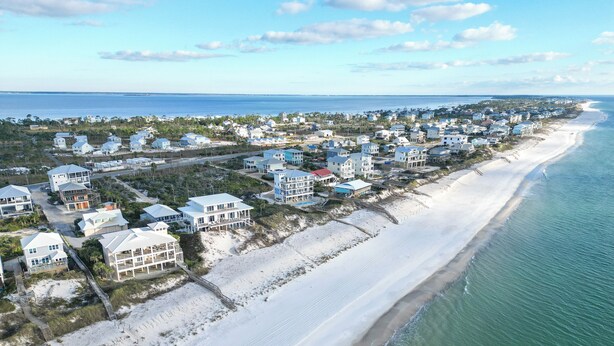 Vue aérienne d’une zone résidentielle côtière, avec des maisons le long de la plage, un ciel dégagé et l’océan en arrière-plan.