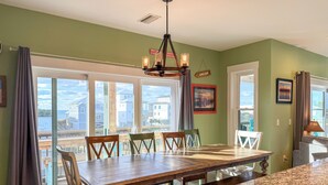 Un coin repas avec une table et des chaises en bois, de grandes fenêtres donnant sur des maisons et un lustre.