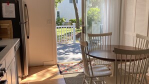 A kitchen with a refrigerator, a dining table with chairs, a patterned rug, and a view of a house and trees through a glass door. - Walk to private beach, central air, wifi, 3 bedrooms. (Chatham)