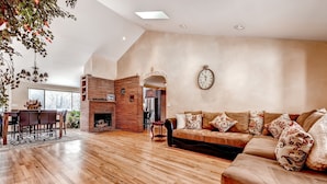 A spacious living room with a wooden floor, a brick fireplace, a large sofa with patterned cushions, and a dining area with a wooden table and chairs. - 3 Bedrooms - Gourmet Kitchen - Open Floorplan - Near Red Rocks & Near Downtown (Lakewood)