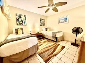A hotel room with two beds, a ceiling fan, artwork on the walls, and a floor fan.