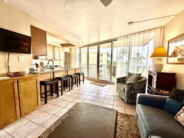 A modern kitchen with a bar area, a dining table, and a sofa in a living area with a view of palm trees.