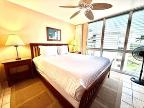 Ein Hotelzimmer mit einem Bett, zwei Nachttischlampen, einem Deckenventilator, einem Fenster mit Jalousien und Blick auf ein Gebäude.