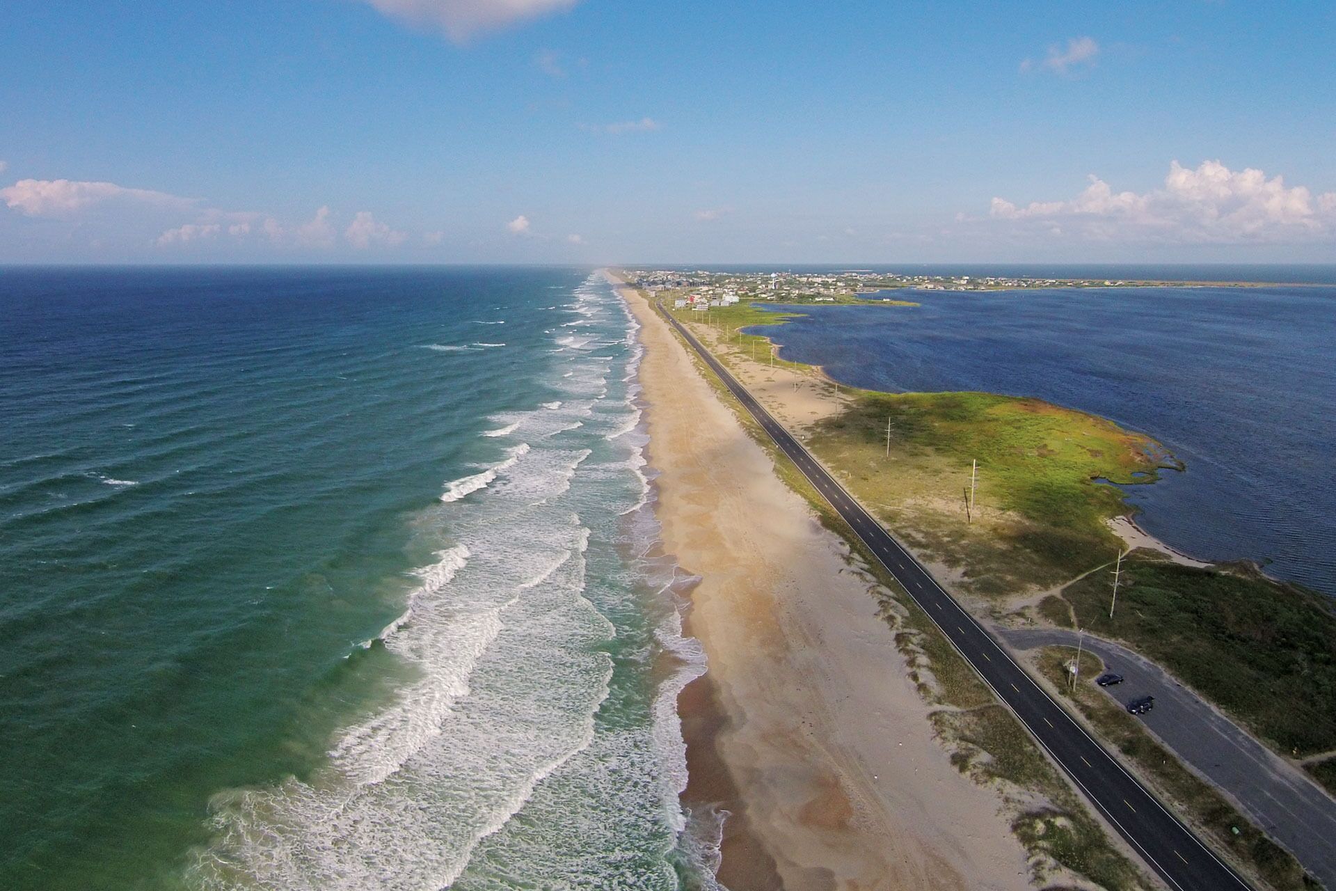 An aerial view of a coastal road with a beach on one side and a cityscape in the distance.