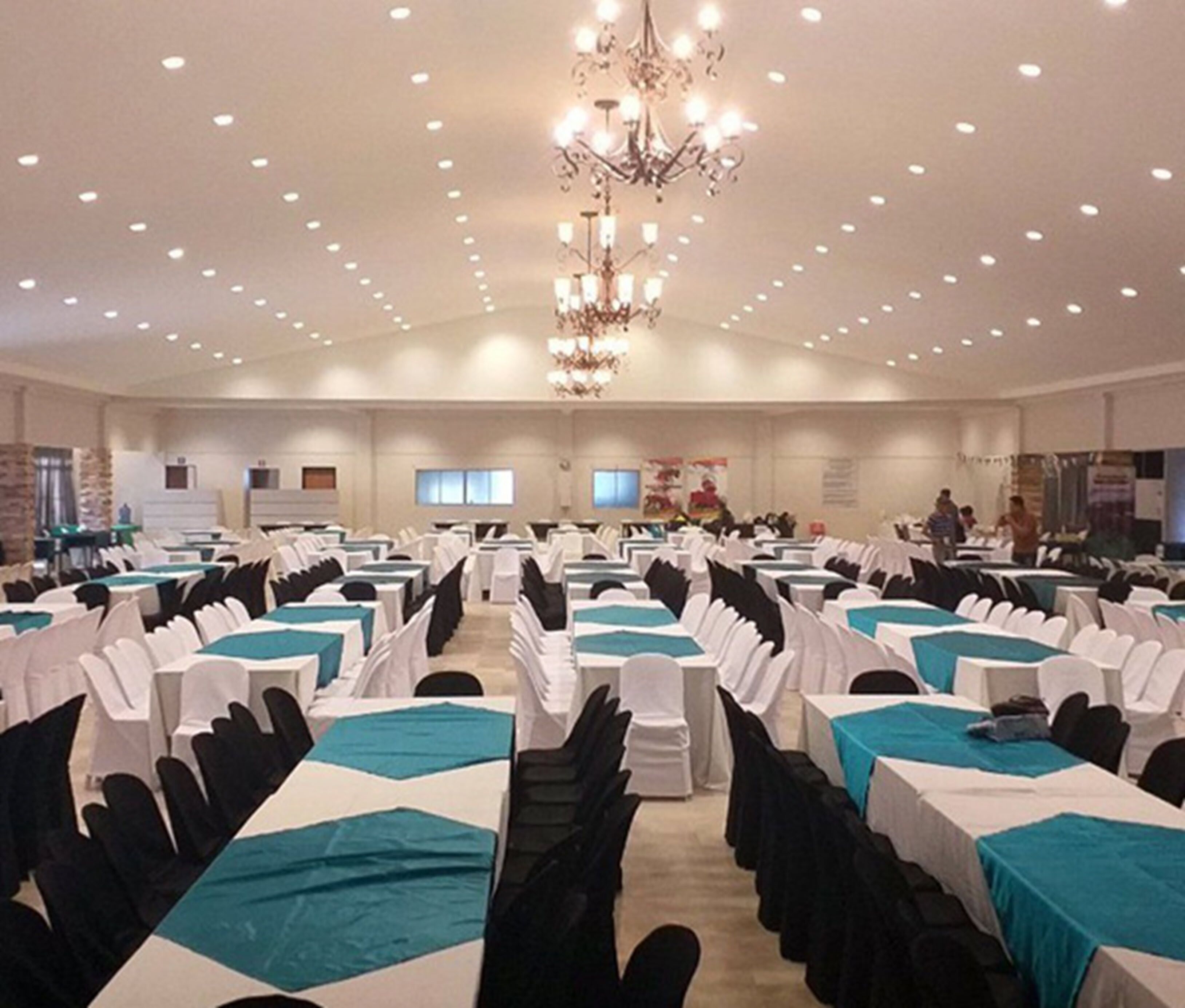 A large banquet hall with rows of tables covered in white tablecloths and teal runners, a chandelier, and recessed ceiling lighting.