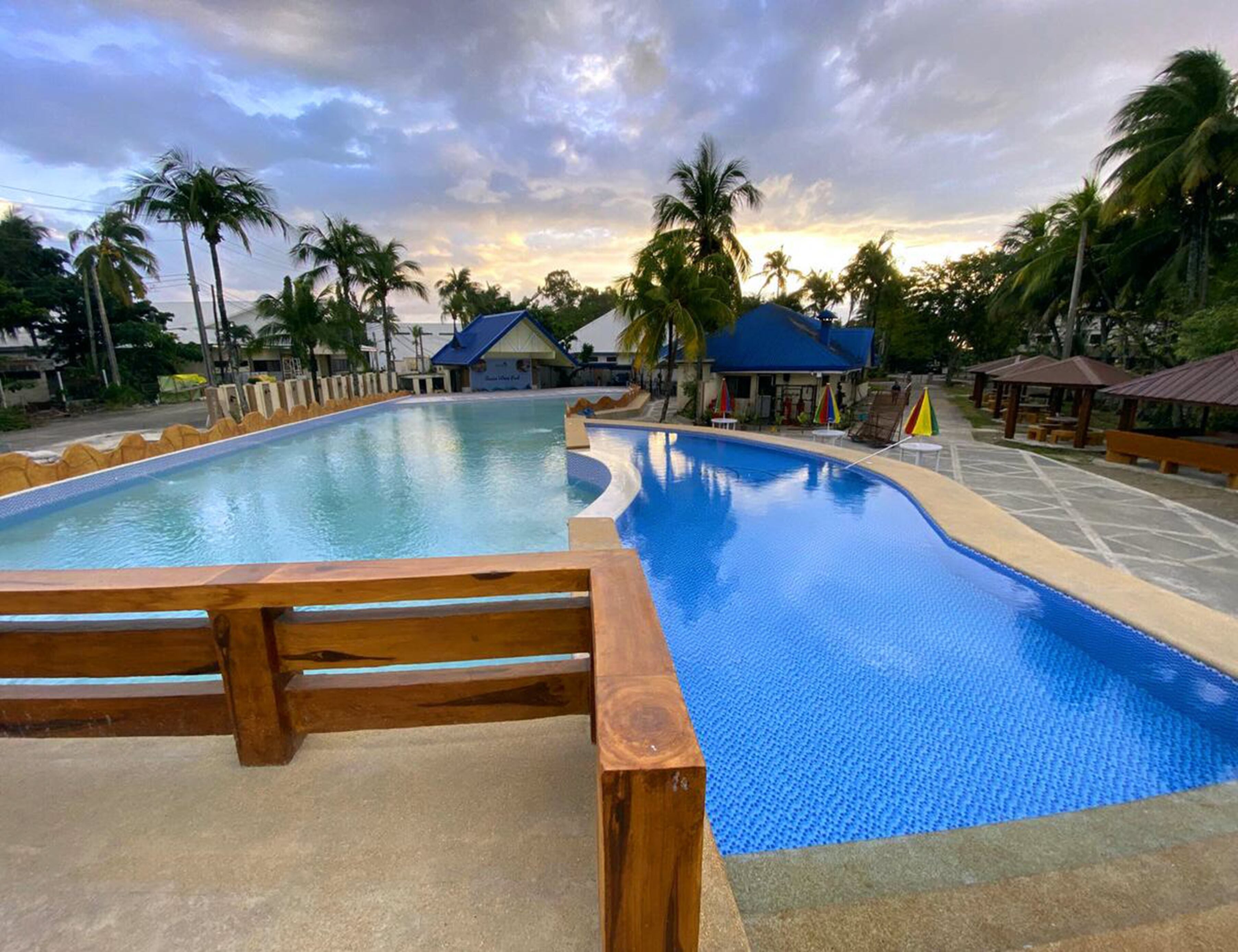 A swimming pool with a wooden deck in the foreground, surrounded by palm trees and small buildings with blue roofs.
