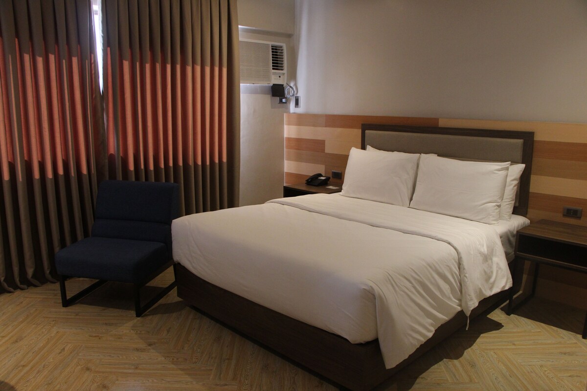 A hotel room with a bed, a chair, a nightstand, and a wall-mounted air conditioning unit.
