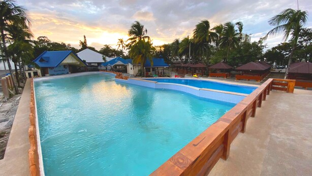 Una gran piscina con muelle de madera y sillas de descanso, rodeada de palmeras y edificios con techos azules.