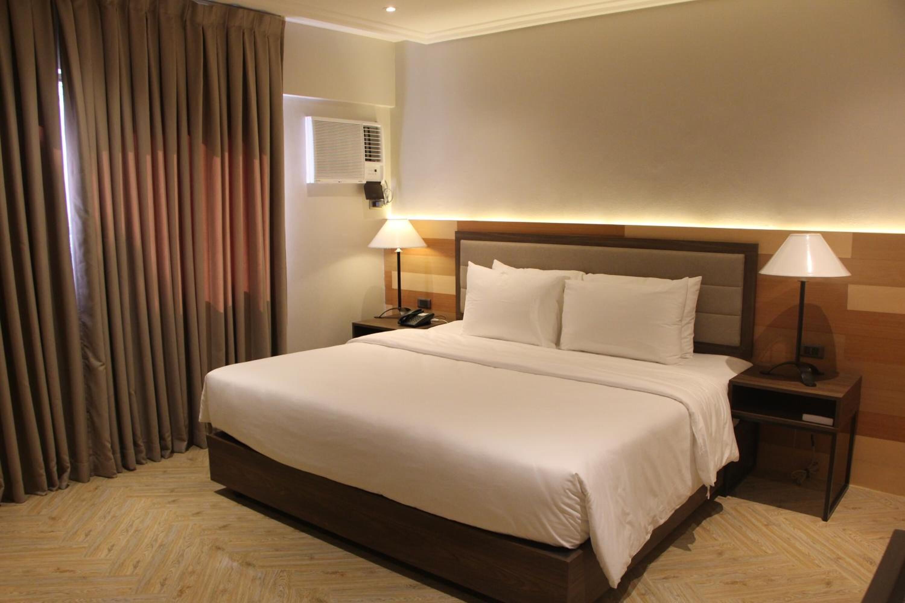 Habitación de hotel con una cama grande, dos mesitas de noche, aire acondicionado en la pared y una ventana con cortinas.