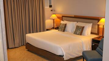 Habitación de hotel con una cama grande, dos mesitas de noche con lámparas, una silla y una ventana con cortinas.