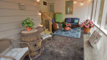 A cozy porch with a wooden bench, a wicker chair, and a cushioned sofa. There are potted plants and a small table with a basket.
