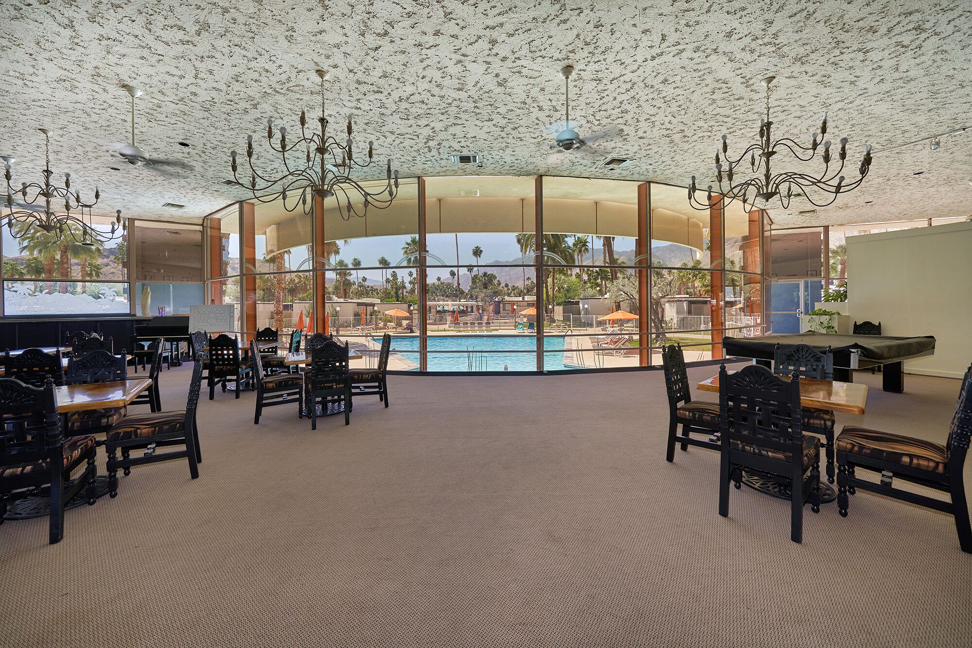 A spacious indoor area with a pool view, featuring a high ceiling with a textured finish, multiple chandeliers, and a seating area with tables and chairs.