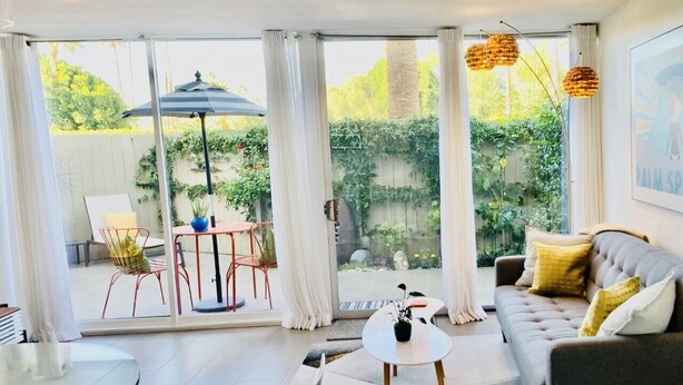 A modern living room with a grey sofa, a round coffee table, and a dining table. There are curtains, a chair, and a small table with a plant.