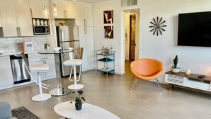 A modern kitchen with stainless steel appliances, a dining area with a white table and orange chairs, and a living room with a grey sofa and a white coffee table.