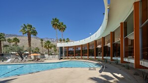 A poolside area with a curved building, palm trees, and a mountain in the background.