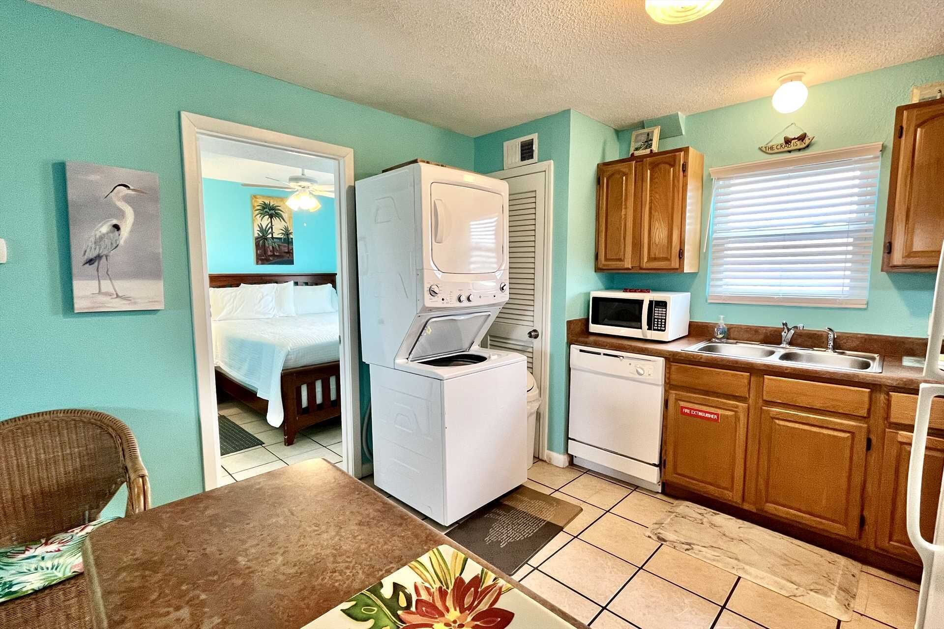 A compact kitchen with a refrigerator, microwave, and dishwasher. There's a dining area with a table and chairs, and a bedroom visible through an open door.