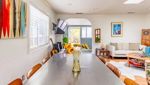 Un comedor con una mesa larga, sillas, un jarrón con girasoles, una guitarra y un televisor en la pared.